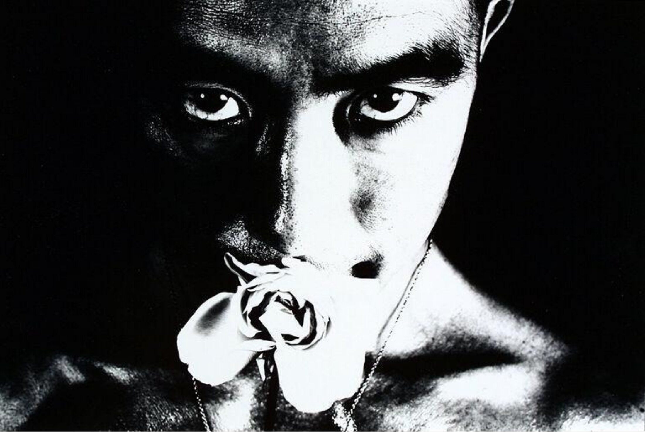 A dramatic black-and-white portrait photograph of a person's face with intense gaze, holding a wilted rose near their mouth against a deep black background.