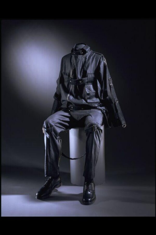 The V&A bondage suit displayed seated, showing the strap running between the knees