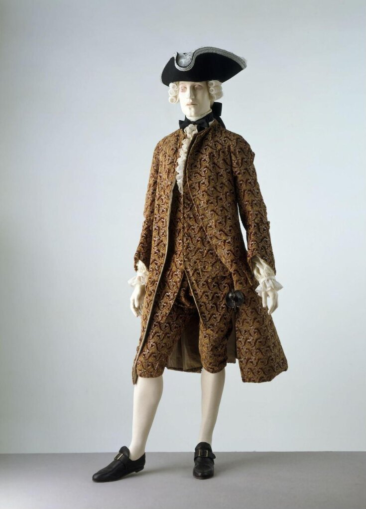 Dandy in semi-formal outfit. He wears a chestnut-brown Savoyard frock coat,  waistcoat, apricot breeches, white hose and buckle shoes. He holds a bicorn  or chapeau bras under his arm. Habit brun Savoyard