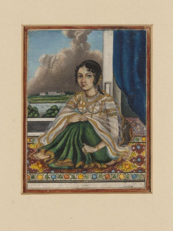 An Indian lady seated on a carpet by an open door overlooking a river with a European-style house on the opposite bank.