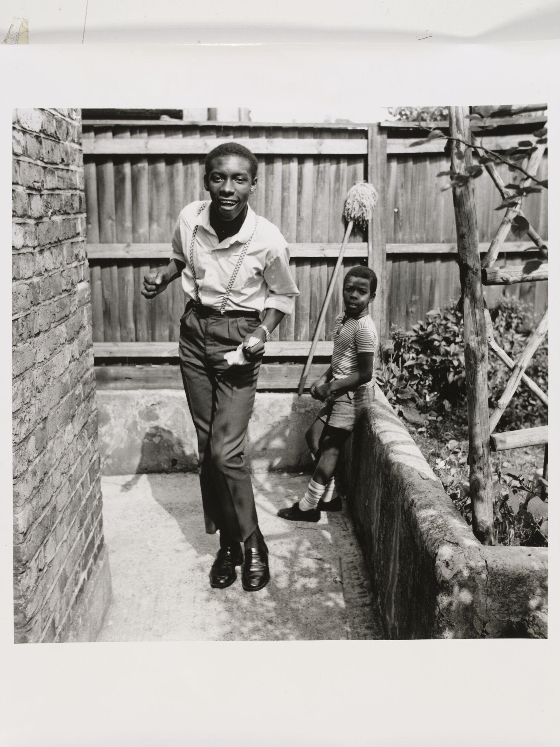 Untitled [Young teenage boy who, just arrived from Jamaica, shows off his latest reggae dance with his younger brother watching, Brixton Hill] by Neil Kenlock