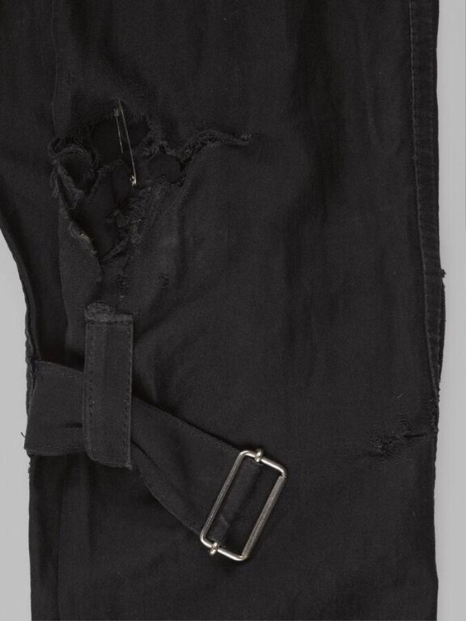 A safety pin spanning a worn-through hole in black cotton bondage trousers